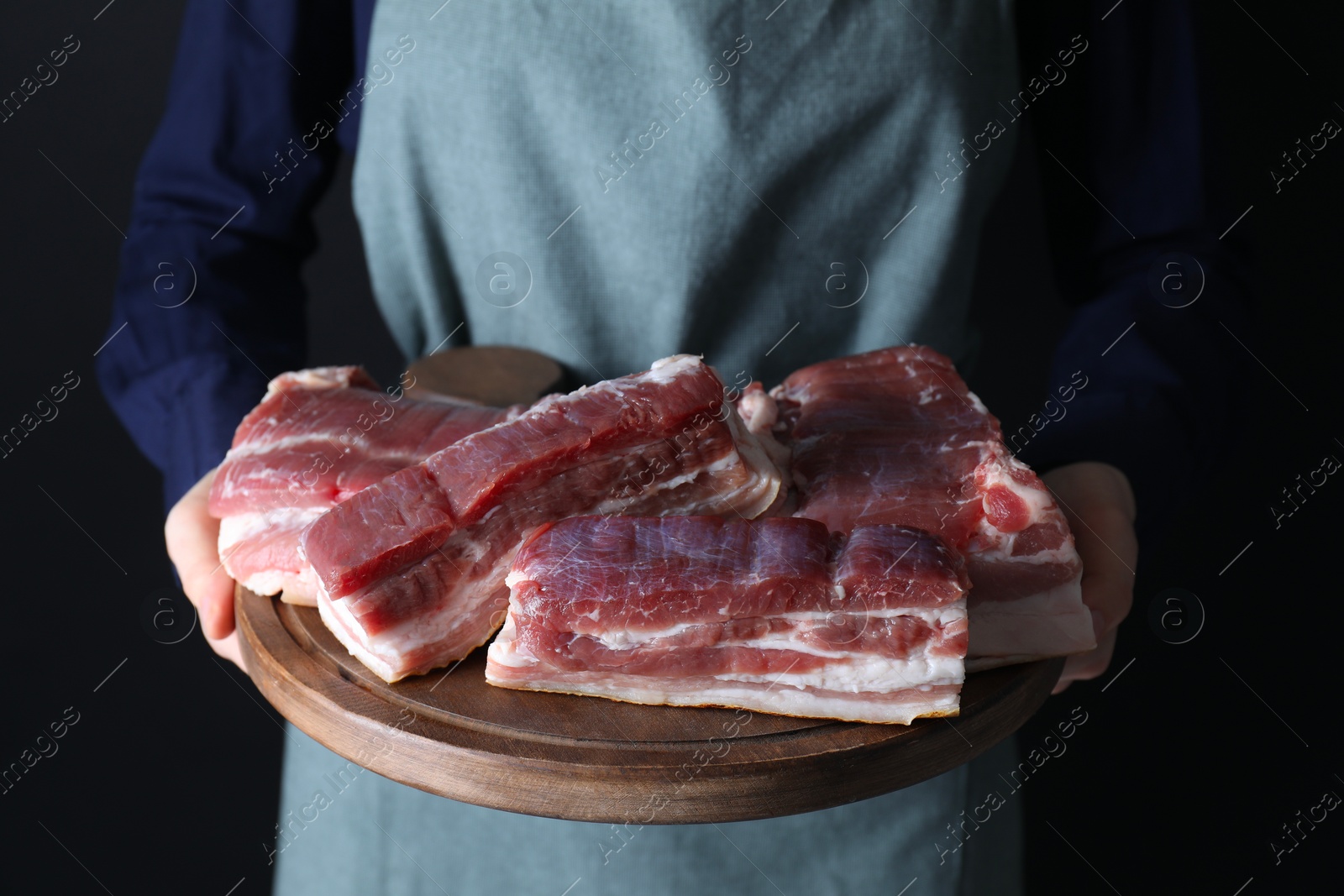 Photo of Woman holding wooden board with pieces of raw pork belly on black background, closeup