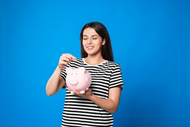 Young woman putting coin into piggy bank on light blue background