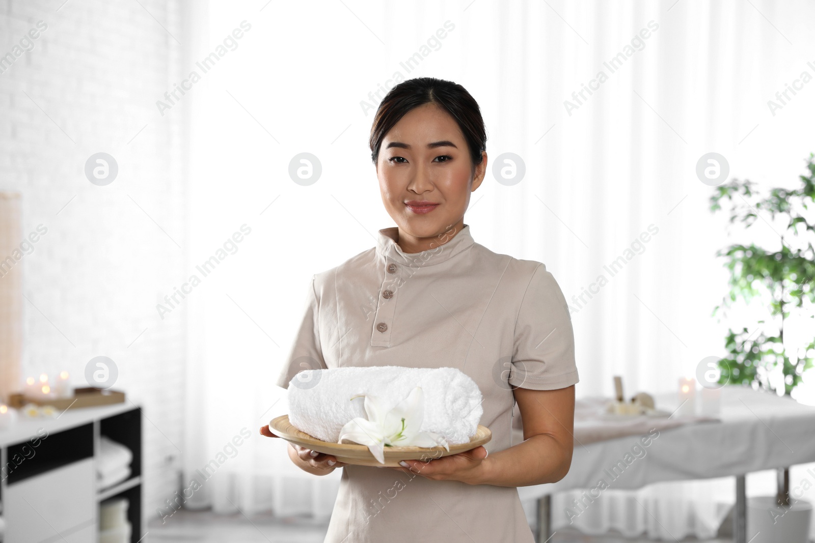 Photo of Portrait of young Asian masseuse holding plate with towel and flower in spa salon