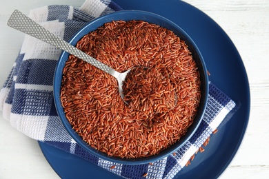 Photo of Bowl with uncooked red rice and spoon on white table, top view