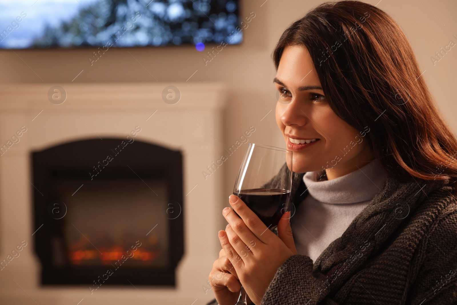 Photo of Young woman with glass of wine resting near fireplace at home