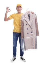 Photo of Dry-cleaning delivery. Happy courier holding coat in plastic bag and showing OK gesture on white background