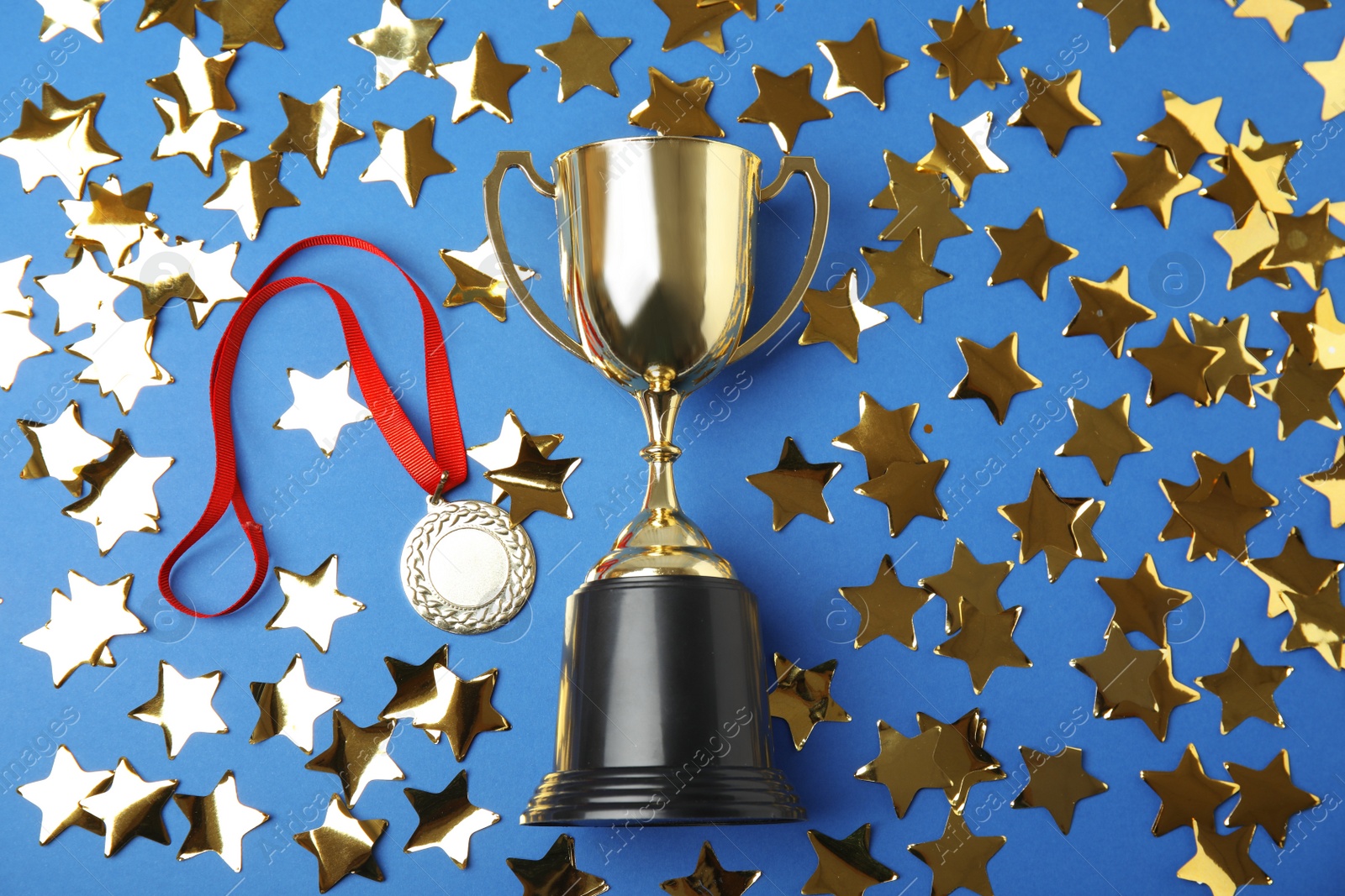 Photo of Gold trophy cup, medal and confetti on blue background, flat lay