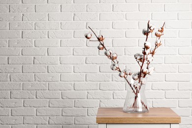 Photo of Cotton branches with fluffy flowers in vase on wooden table near white brick wall. Space for text