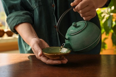 Photo of Woman pouring freshly brewed tea from teapot into cup at wooden table indoors, closeup. Traditional ceremony
