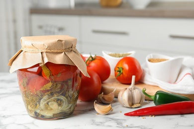Photo of Pickled tomatoes in glass jar and products on white marble table in kitchen