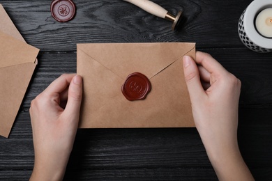 Photo of Woman holding envelope with wax seal at black wooden table, top view