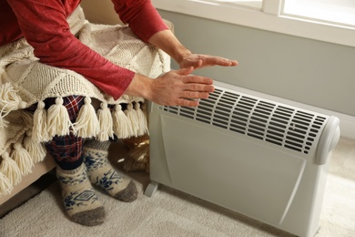 Man warming hands near electric heater at home, closeup