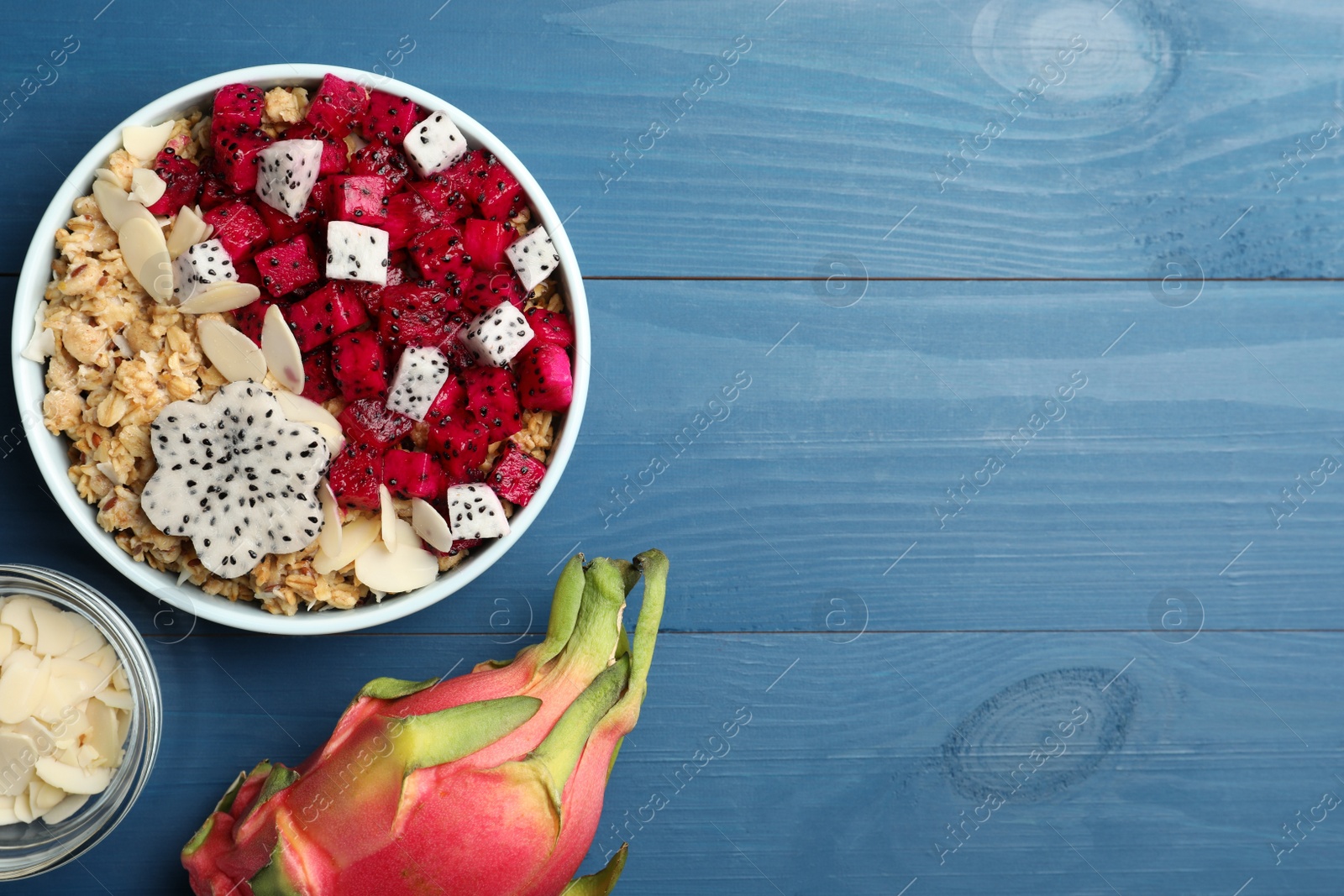 Photo of Bowl of granola with pitahaya and almond petals on blue wooden table, flat lay. Space for text