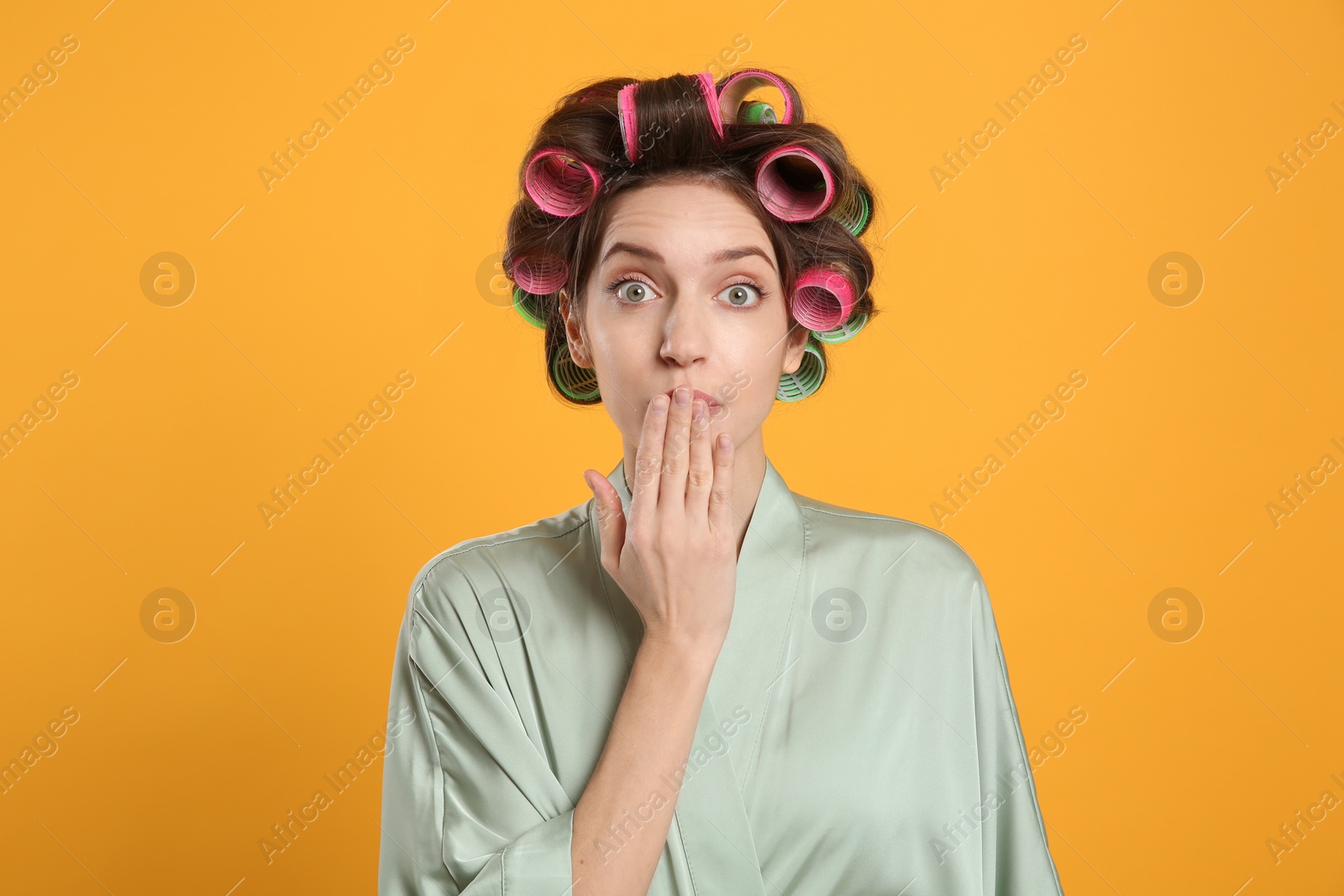 Photo of Emotional young woman in silk bathrobe with hair curlers on yellow background