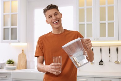 Photo of Happy man pouring water from filter jug into glass in kitchen