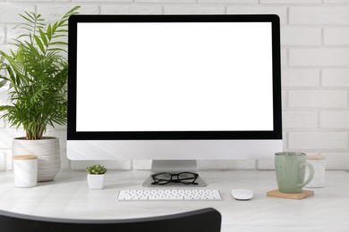 Photo of Office workplace with computer, glasses, cup and houseplants on light table near white brick wall