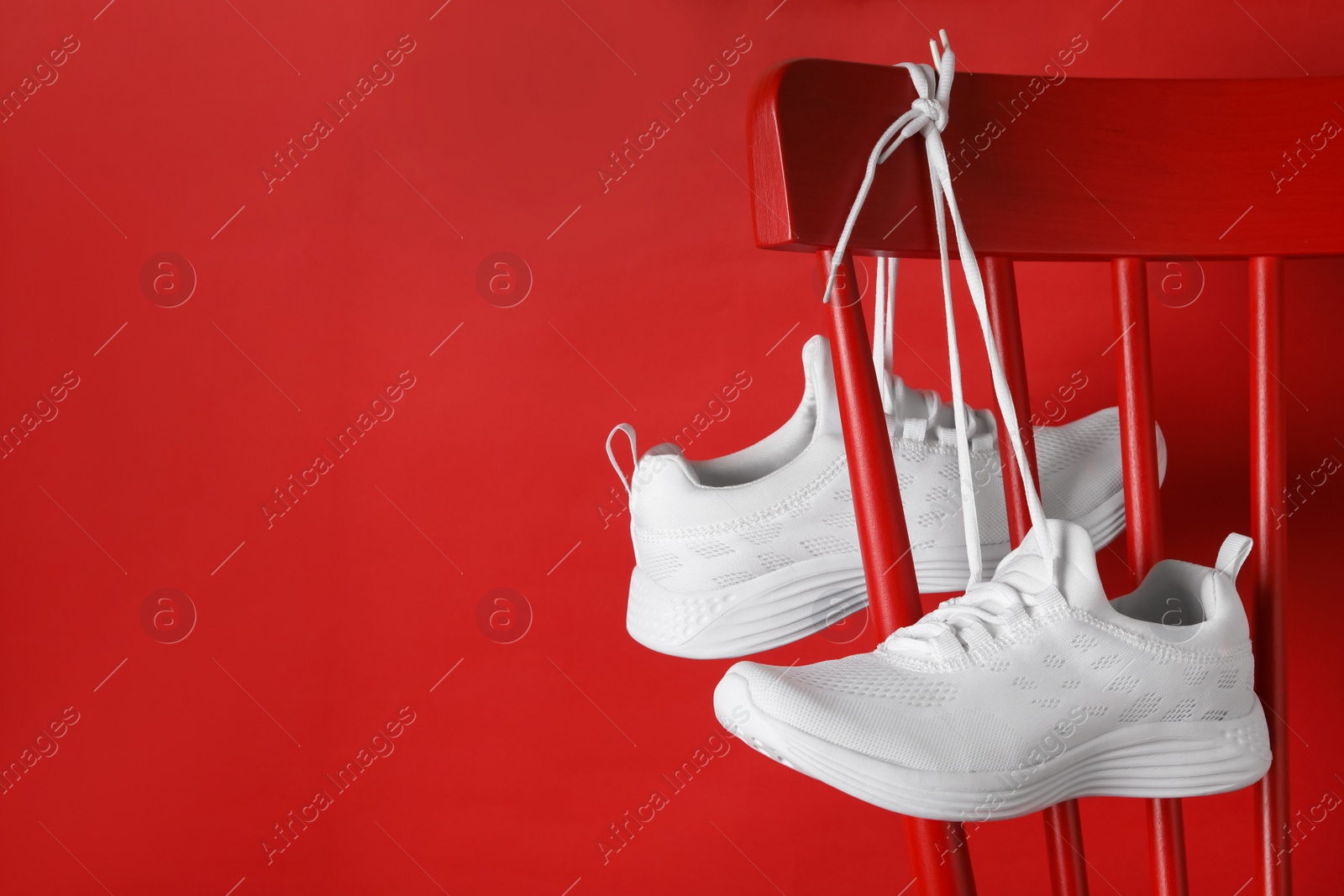 Photo of Stylish sneakers with white shoe laces hanging on chair against red background. Space for text
