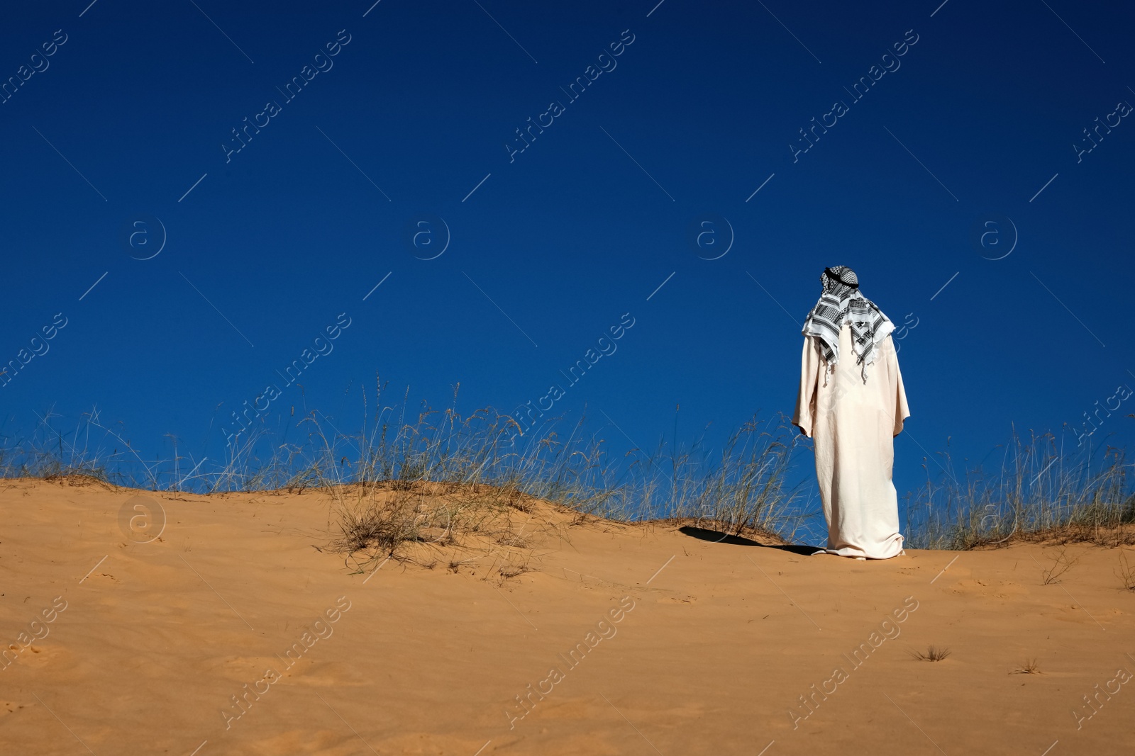 Photo of Man in arabic clothes walking through desert on sunny day, back view