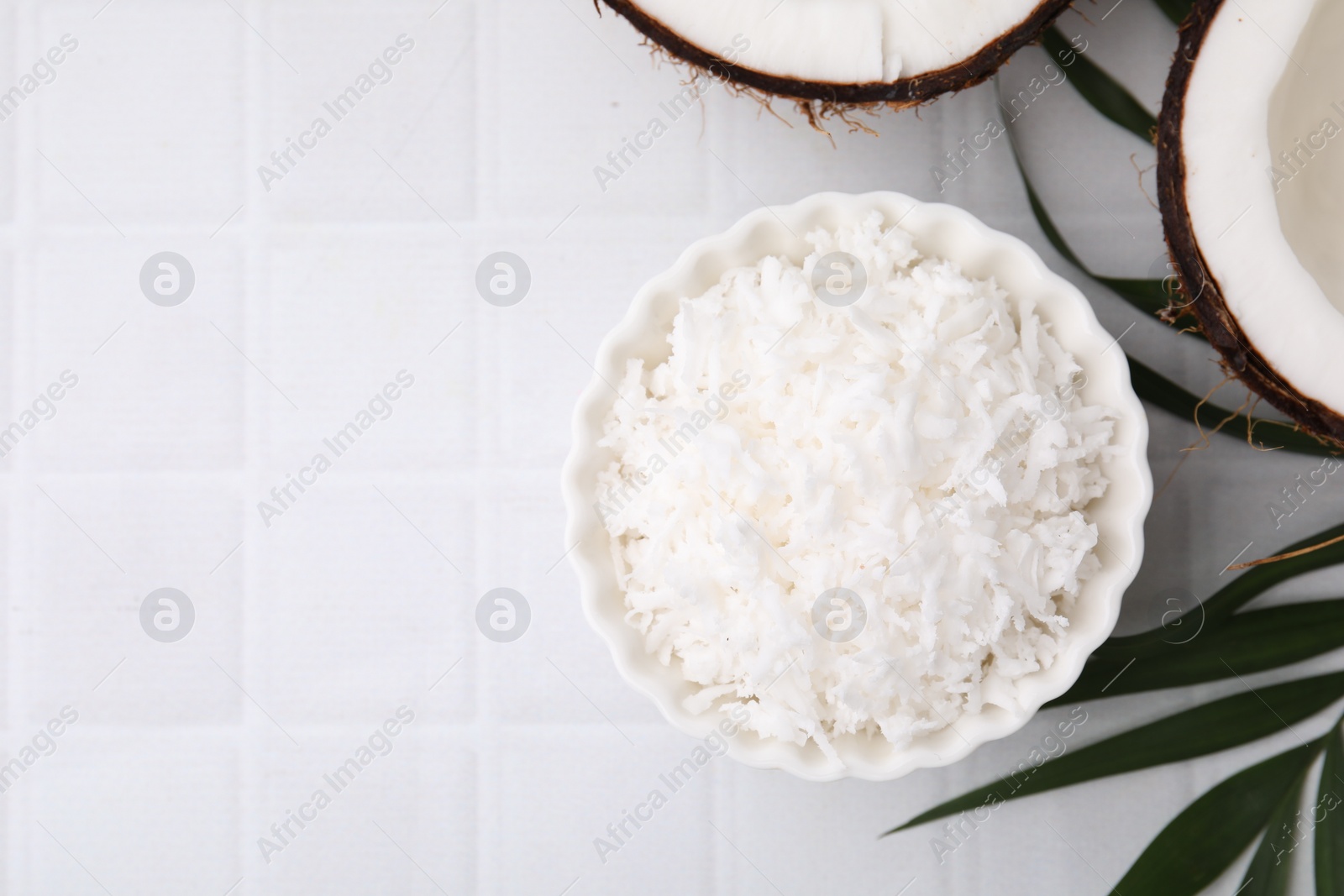 Photo of Coconut flakes in bowl, nuts and palm leaf on white tiled table, flat lay. Space for text