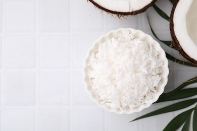 Coconut flakes in bowl, nuts and palm leaf on white tiled table, flat lay. Space for text