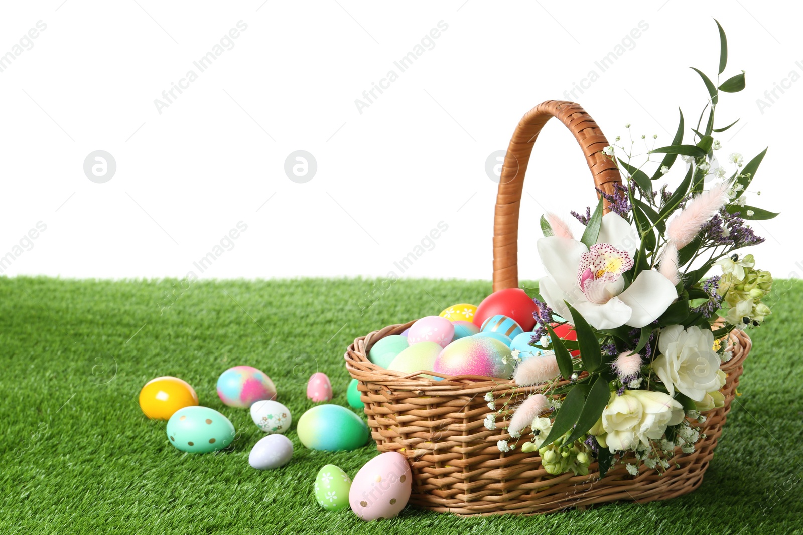 Photo of Wicker basket with Easter eggs and beautiful flowers on green grass against white background. Space for text