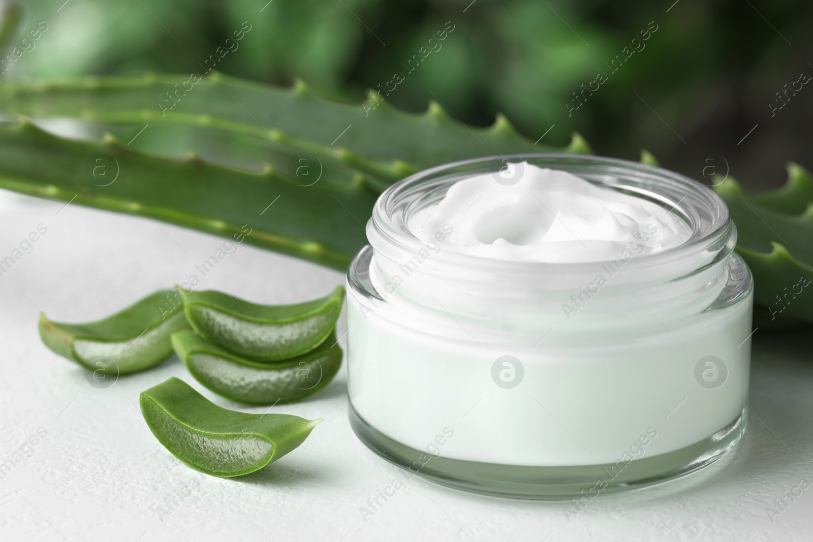 Photo of Jar with cream and cut aloe leaves on white table against blurred green background, closeup