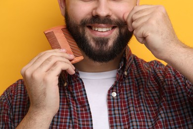 Photo of Handsome young man combing beard on yellow background, closeup