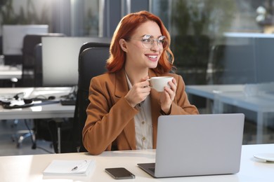 Photo of Happy woman with cup of drink working on laptop at white desk in office