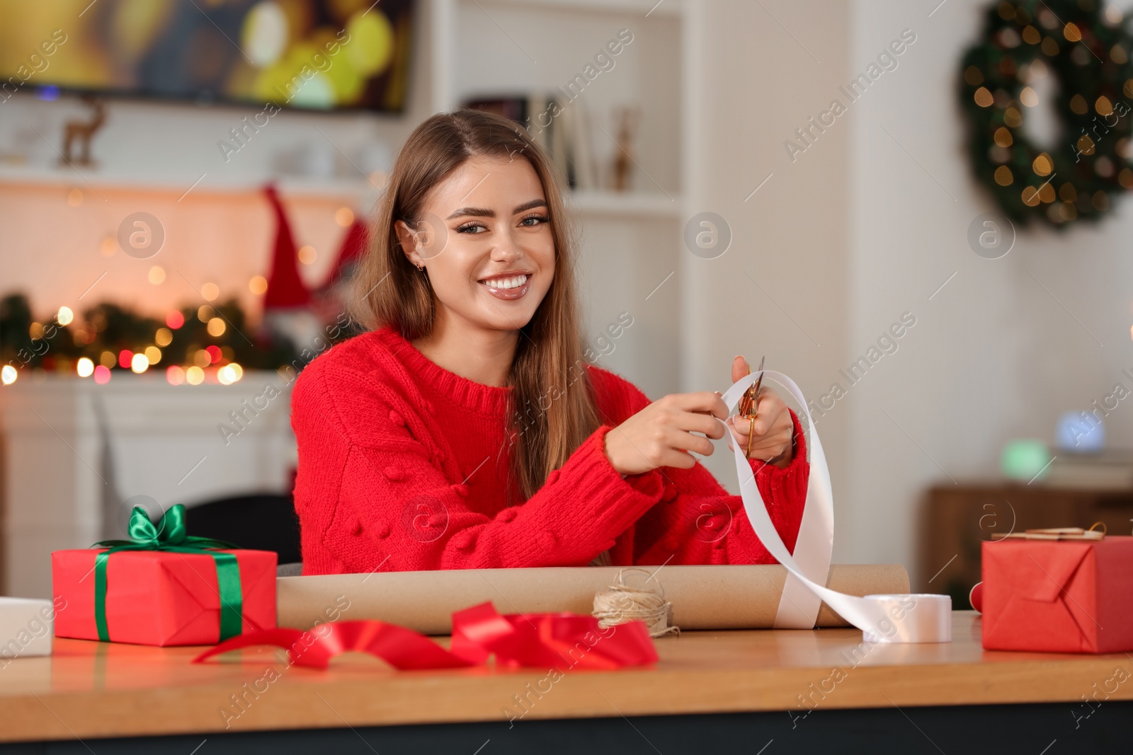 Photo of Beautiful young woman with white ribbon at table in room. Decorating Christmas gift