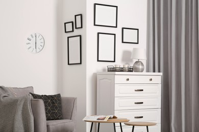 Photo of Empty frames hanging on white wall, chest of drawers and wooden tables indoors