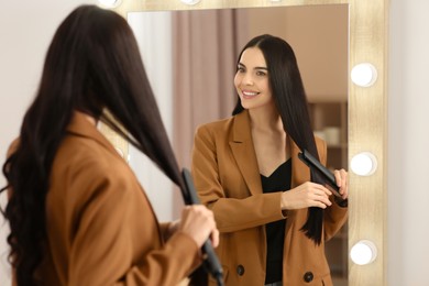 Photo of Beautiful happy woman using hair iron near mirror in room