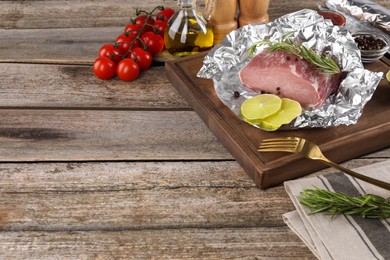 Photo of Aluminum foil with raw meat, rosemary, lime and spices on wooden table, space for text