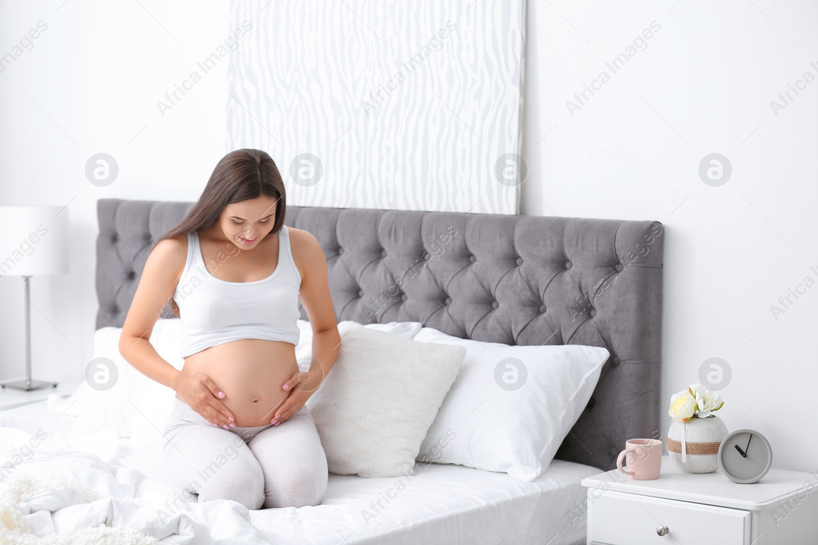 Photo of Young beautiful pregnant woman sitting on bed and touching her belly at home