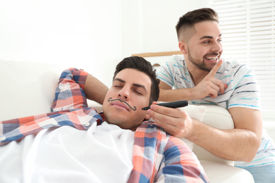 Photo of Young man drawing mustache on face of sleeping friend indoors. April fool's day