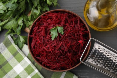 Grated red beet with parsley in bowl, grater and oil on table, flat lay