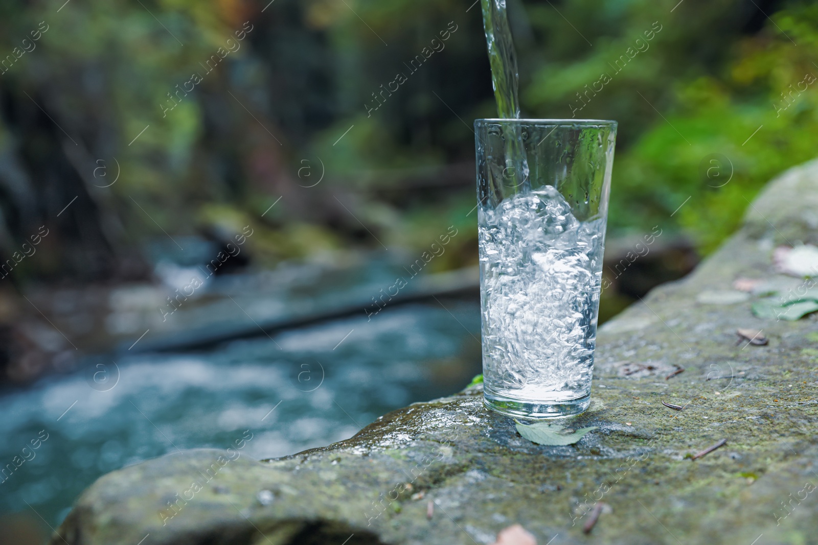 Photo of Fresh water pouring into glass on stone near stream. Space for text