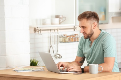 Photo of Young handsome man working with laptop at table in kitchen