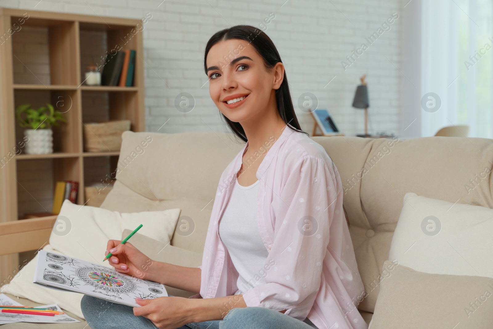 Photo of Young woman coloring antistress page on sofa in living room