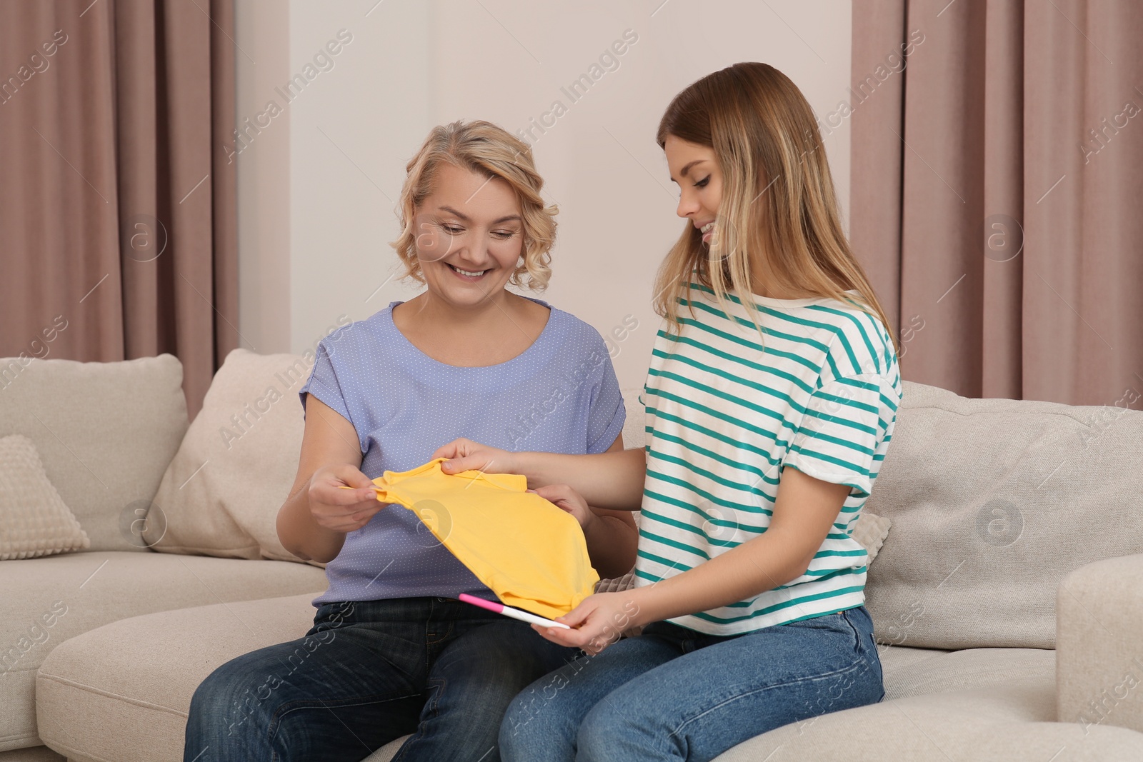 Photo of Young woman showing her mother baby's bodysuit and pregnancy test at home. Grandparent reaction to future grandson