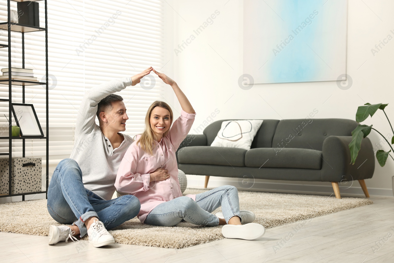 Photo of Young family housing concept. Pregnant woman with her husband forming roof with their hands while sitting on floor at home