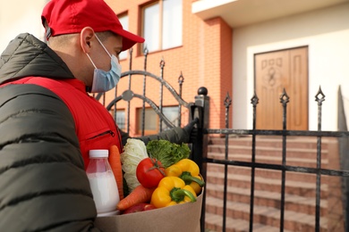 Courier in medical mask holding paper bag with groceries and ringing gate bell outdoors. Delivery service during quarantine due to Covid-19 outbreak