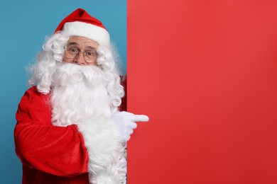 Photo of Santa Claus pointing at blank red poster on light blue background, space for text