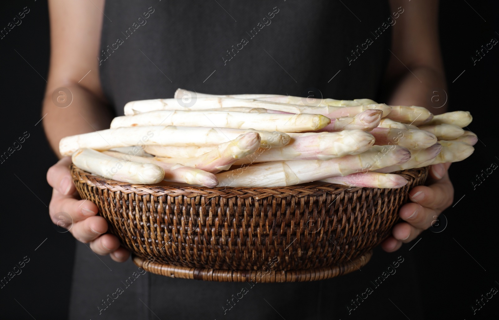 Photo of Woman holding wicker bowl with fresh white asparagus on black background, closeup