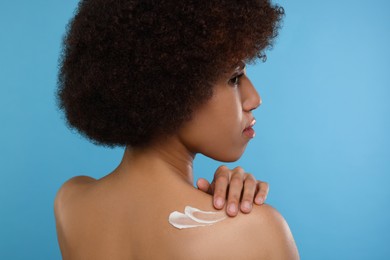 Photo of Beautiful young woman applying body cream onto back on light blue background