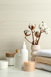 Photo of Bath accessories. Personal care products and cotton flowers in vase on white marble table near light wooden wall