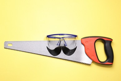 Photo of Man's face made of artificial mustache, safety glasses and hand saw on yellow background, top view