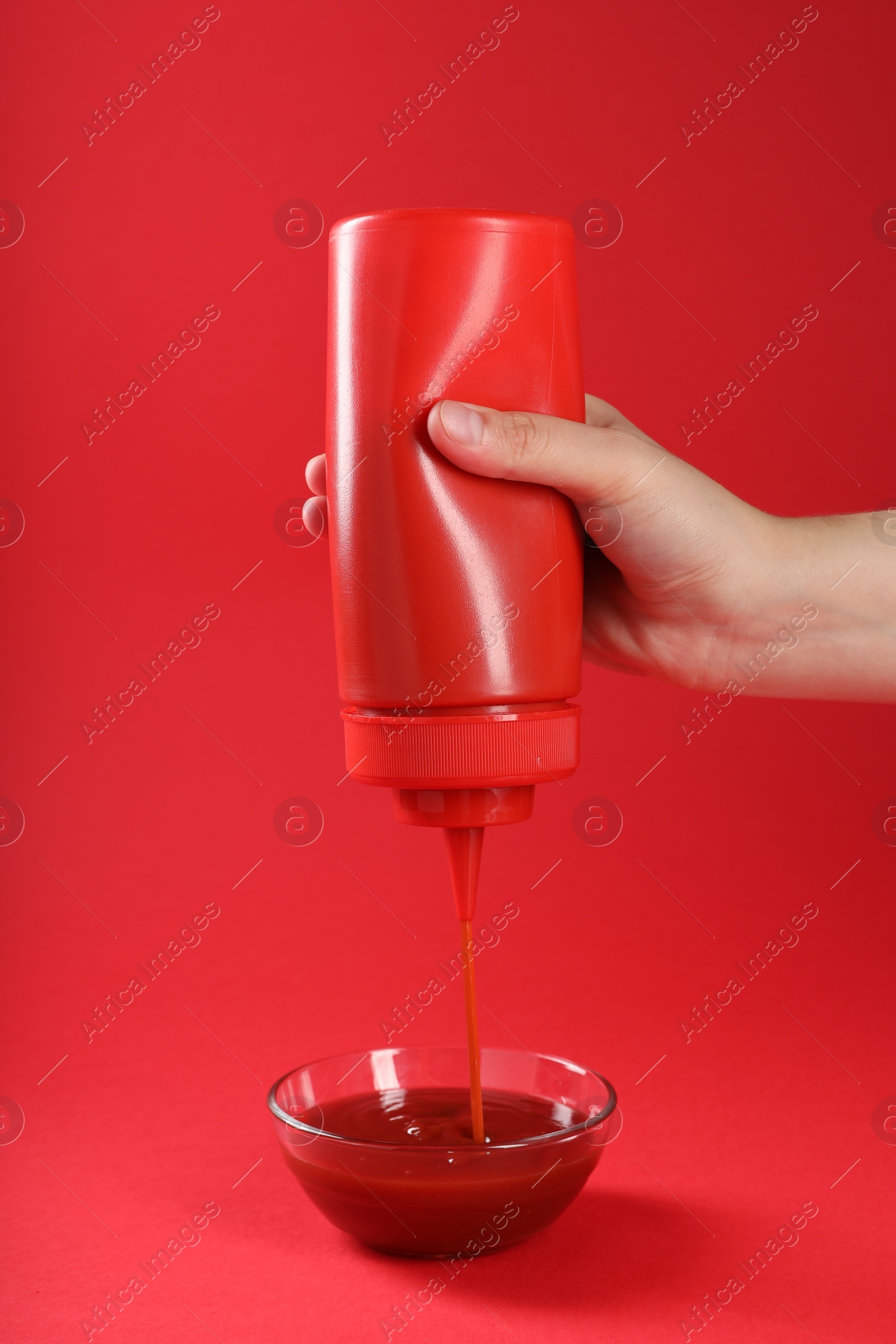 Photo of Woman pouring tasty ketchup from bottle into bowl on red background, closeup