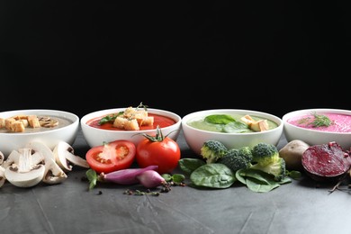 Photo of Different tasty cream soups in bowls and ingredients on dark grey table