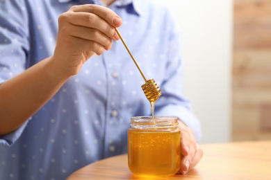 Photo of Woman holding honey dipper over jar at wooden table, closeup