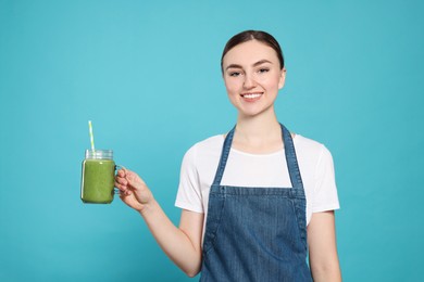 Beautiful young woman in denim apron with delicious smoothie on light blue background