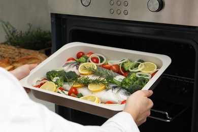 Photo of Woman putting baking dish with raw fish and vegetables into oven in kitchen, closeup