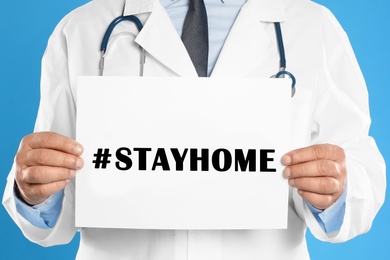 Senior doctor holding paper with hashtag Stayhome on light blue background, closeup. Protective measure during coronavirus pandemic