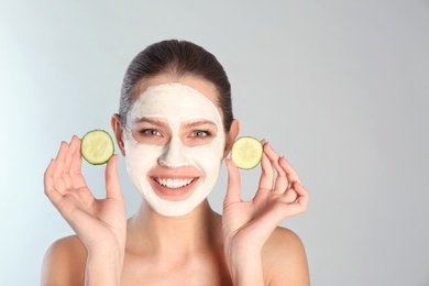 Photo of Beautiful woman holding cucumber slices near her face with clay mask against grey background. Space for text