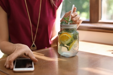 Photo of Young woman with mason jar of tasty natural lemonade and mobile phone on table in cafe, closeup. Detox drink